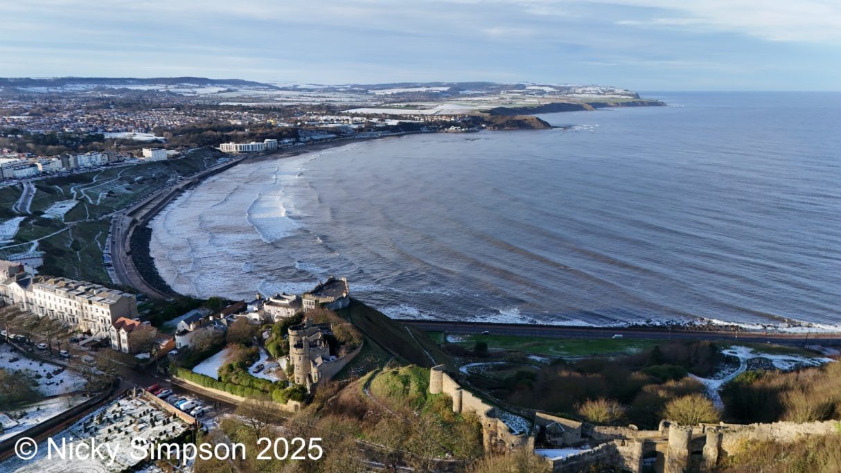 Panoramic view of a coastal landscape with a beach, waves, and distant hills.