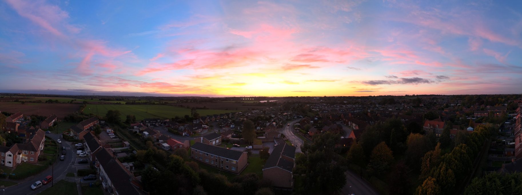 Panoramic view of a sunset over a rural landscape with scattered houses and vibrant clouds.