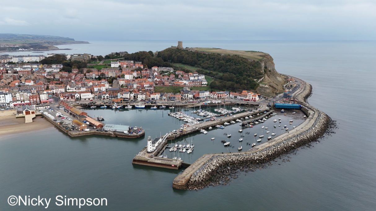 Aerial view of a coastal village with a marina and a hilltop castle in the background.
