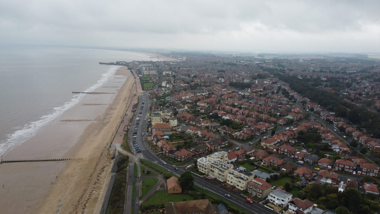 Aerial view of a coastal town with beach, sea, and residential streets under a cloudy sky.