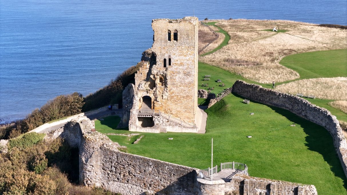 Ruined castle on a cliff overlooking a coastline and grassy landscape.