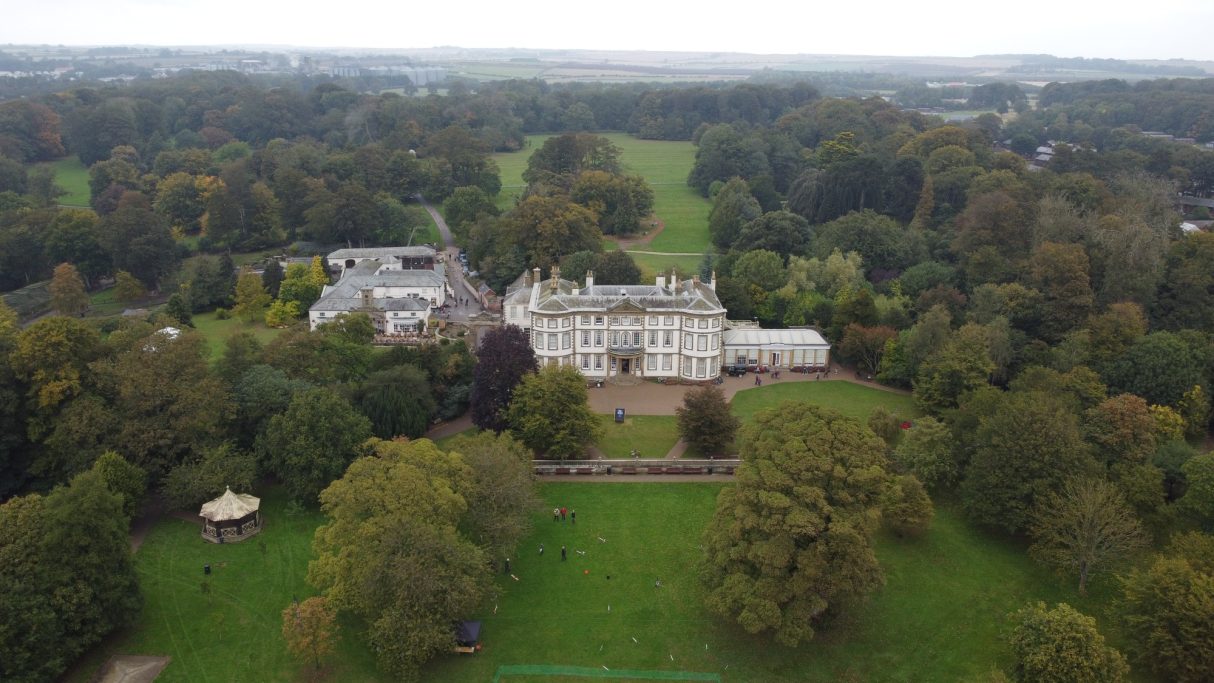 Aerial view of a grand mansion surrounded by lush green trees and open parkland.