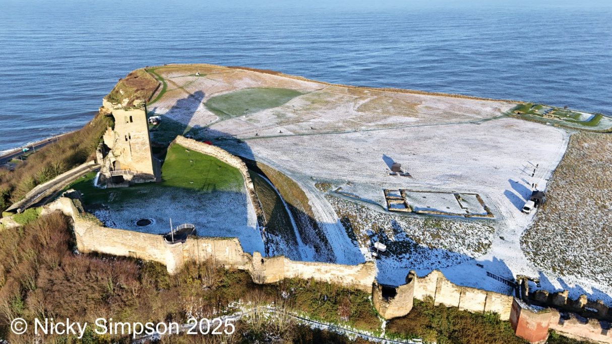 Aerial view of a coastal castle with ruins and green fields beside the sea.