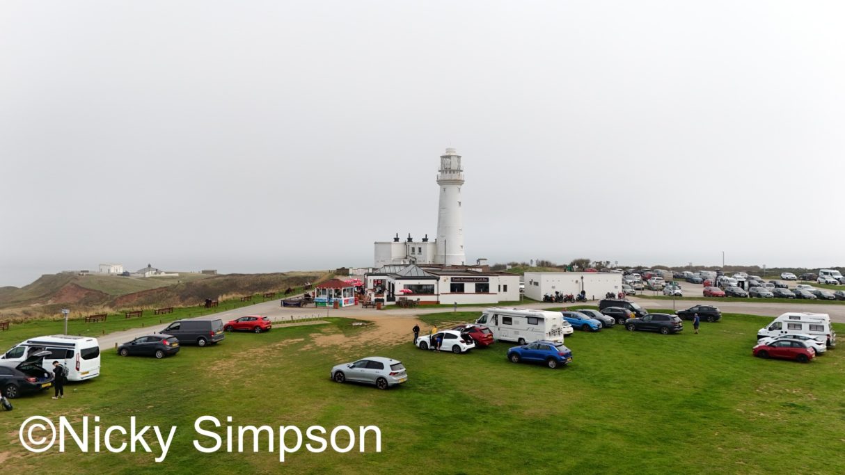 Coastal scene with a lighthouse and a parking area filled with cars.