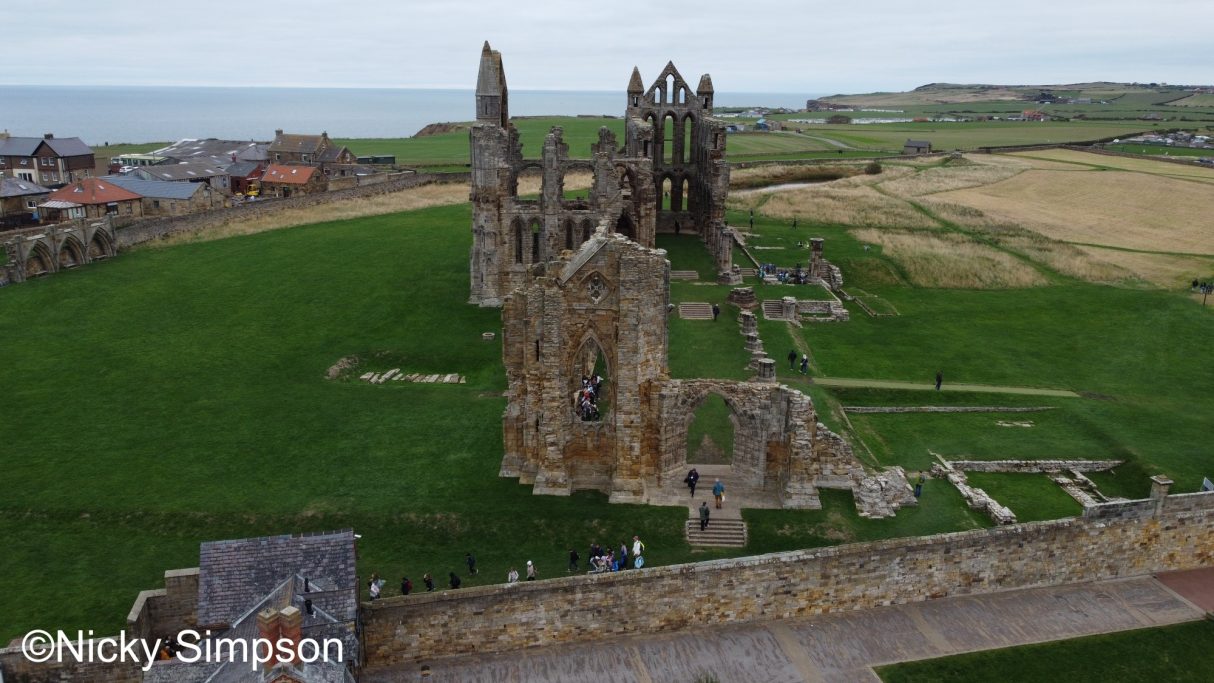 Ruins of a medieval abbey surrounded by green fields and distant coastline.