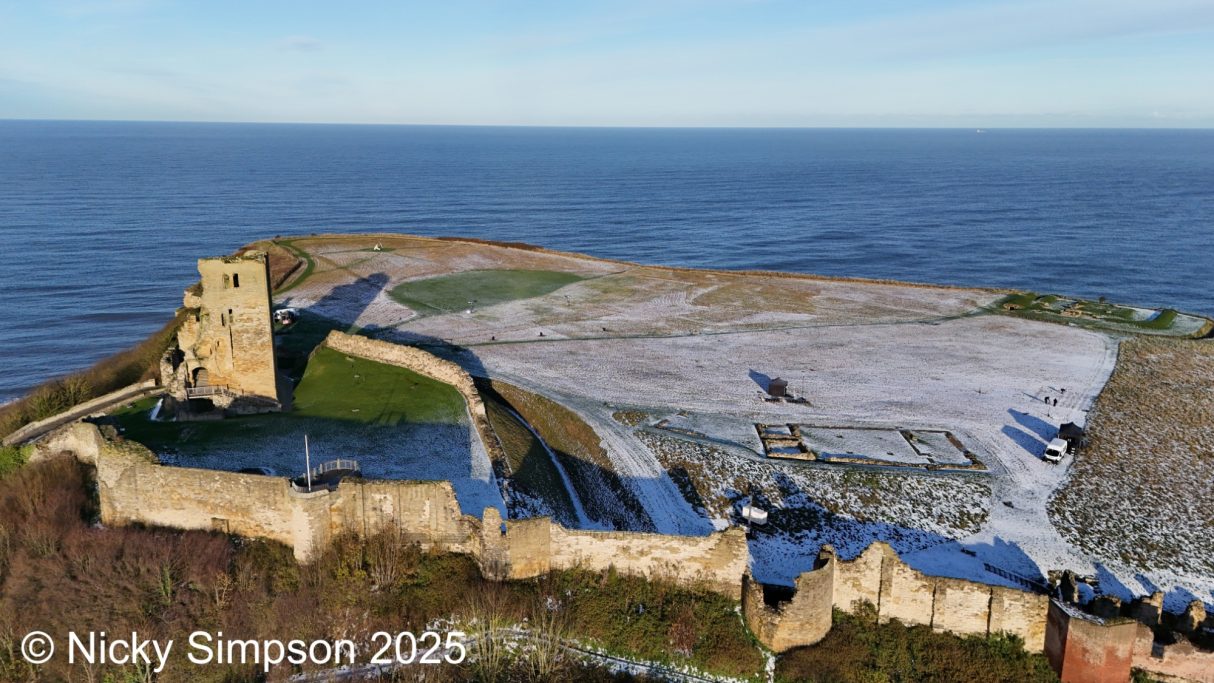 Aerial view of a coastal castle ruins with snow on the ground and the sea in the background.