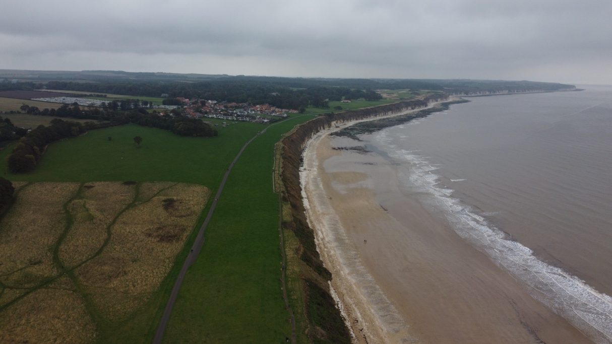 Coastal landscape with cliffs, sandy beach, and green fields under a cloudy sky.