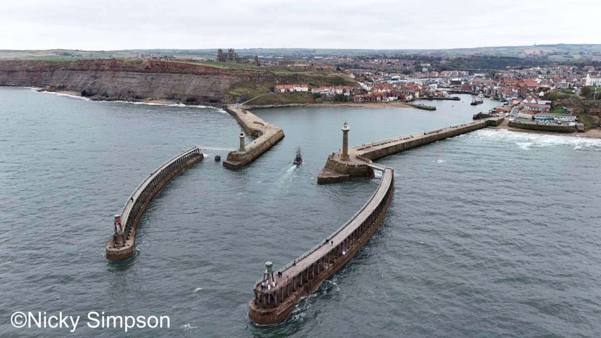 Aerial view of a coastal harbour with stone piers and a distant town.