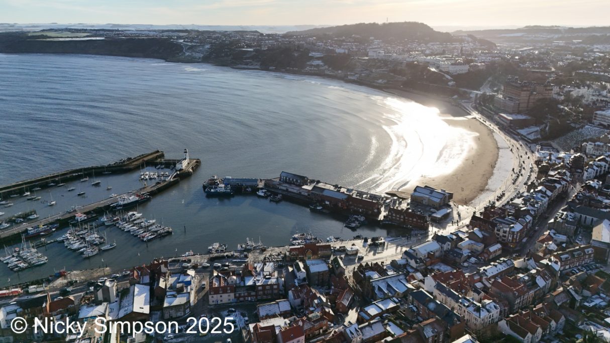 Aerial view of a coastal town with marina, beach, and sunlit hills in the background.