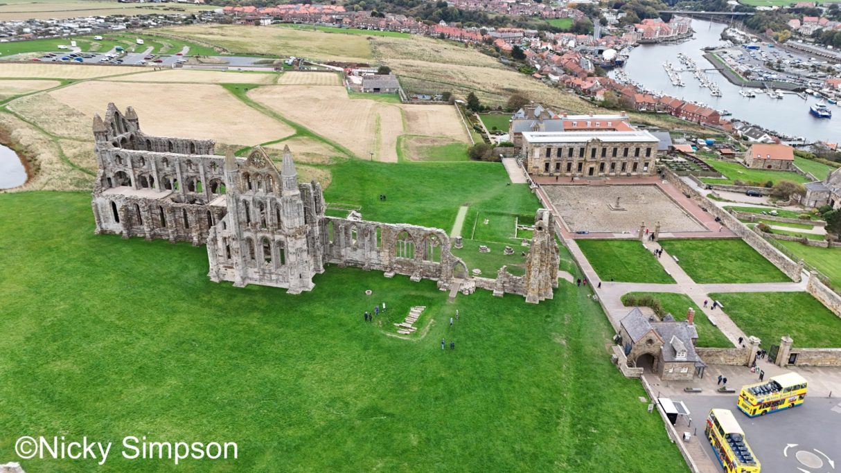 Aerial view of a ruined abbey alongside a formal building and lush green landscape.